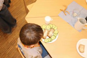 Kind sitzt zum Frühstück am Tisch mit einem Teller mit Gurken und einer Scheibe Brot mit Frischkäse